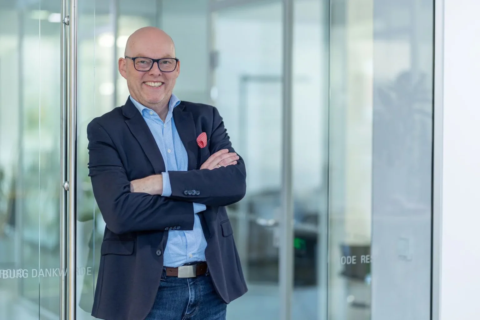 Lächelnder Geschäftsführer Martin Streppel mit Brille, marineblauem Blazer, blauem Hemd und Jeans steht mit verschränkten Armen in einem modernen Büro mit Glaswänden.